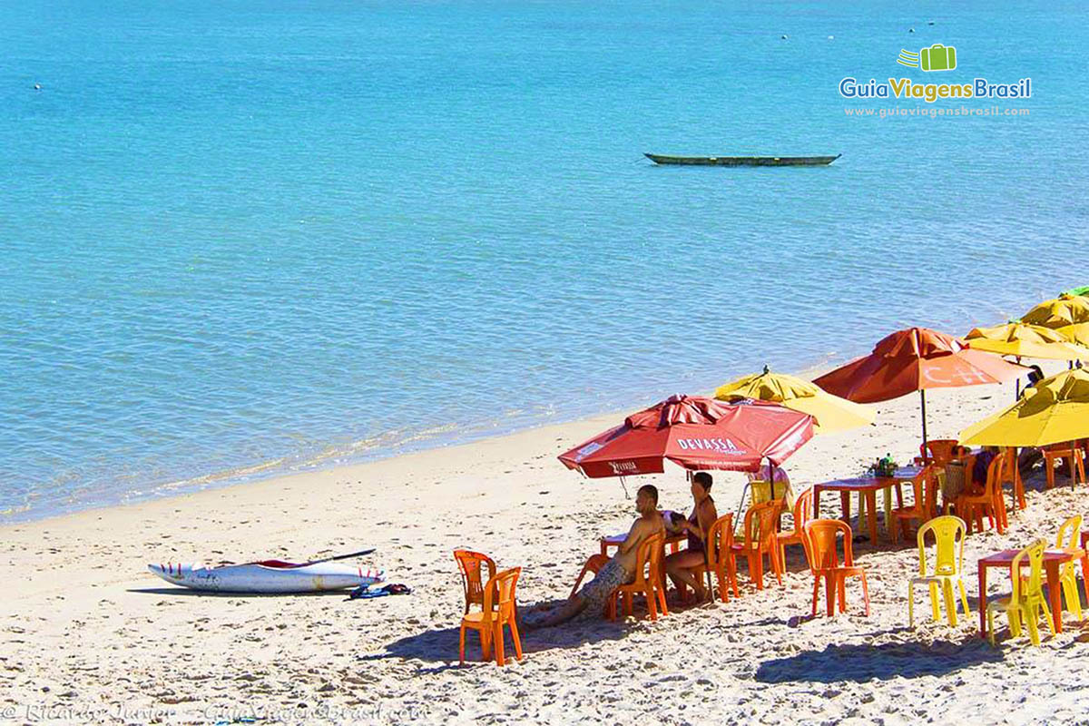 Imagem de casal de turistas sentados na praia e uma caiaque na areia.