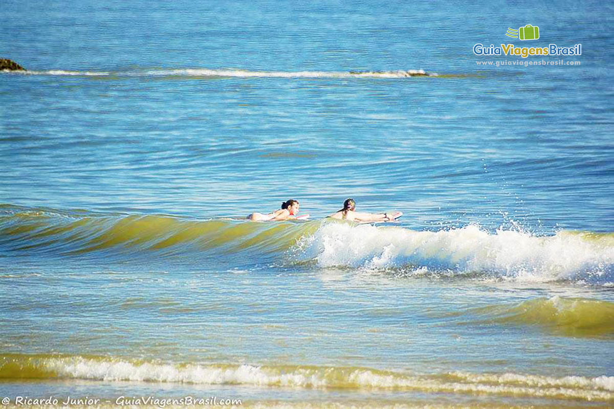 Imagem de duas meninas entrando no mar com as pranchas para surfar, na Praia da Barra do Sai, em Itapoá, Santa Catarina, Brasil.