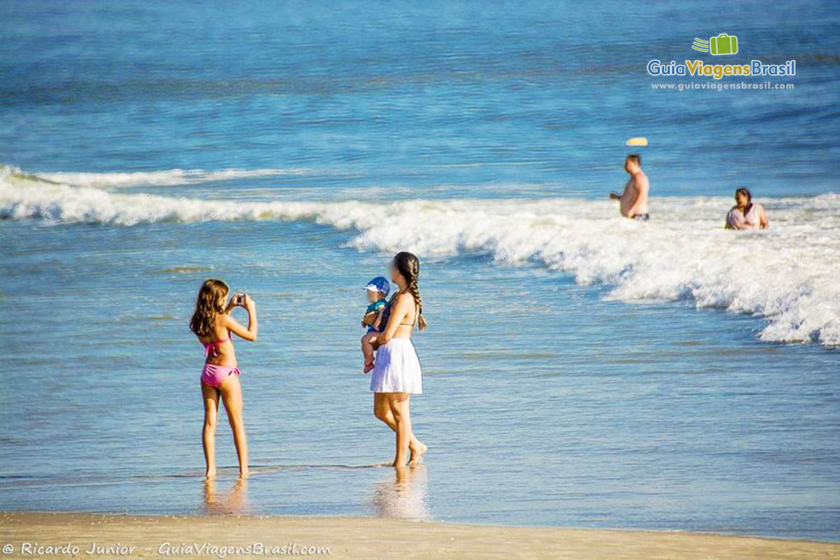 Imagem de uma moça com bebê no colo e uma criança tirando fotos deles na beira do mar, na Praia da Barra do Sai, em Itapoá, Santa Catarina, Brasil.