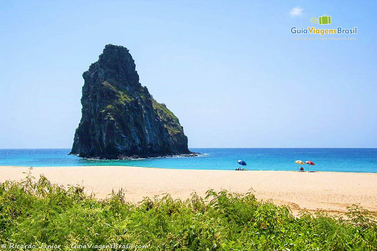 Imagem de três guarda sol na areia e no mar uma rocha, compondo a belíssima paisagem, da Praia da Cacimba do Padre, em Fernando de Noronha, Pernambuco, Brasil.