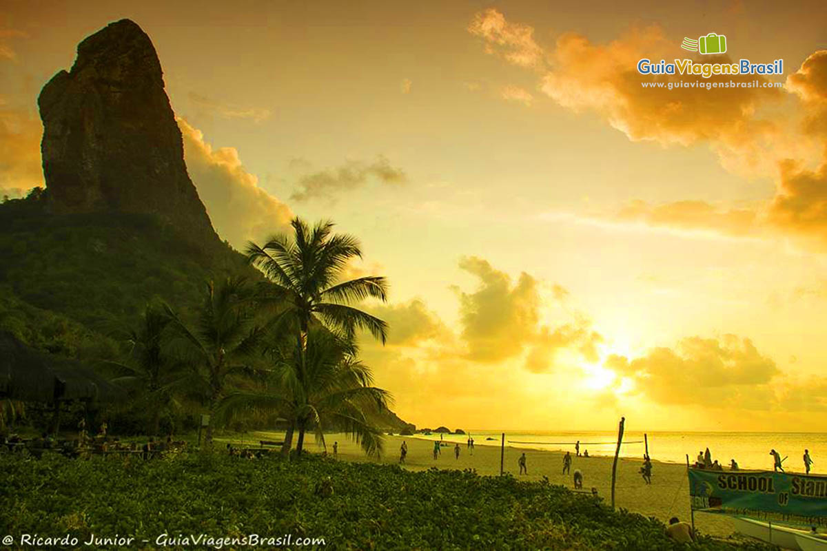 Imagem de uma Pedra alta no lado esquerdo e pessoas jogando vôlei no entardecer, na Praia da Conceição, em Fernando de Noronha, Pernambuco, Brasil.