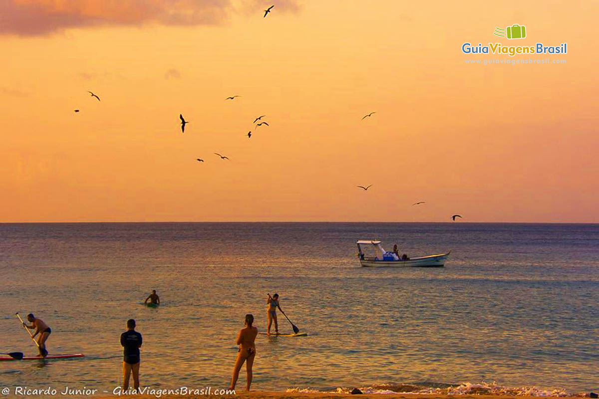 Imagem de fim de tarde com turistas no mar praticando stand up, barco de pescador no mar e gaivotas o céu, na Praia da Conceição, em Fernando de Noronha, Pernambuco, Brasil.