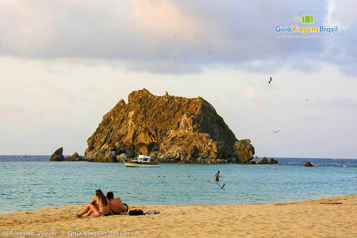 Imagem de casal na areia admirando a linda paisagem da Praia da Conceição, em Fernando de Noronha, Pernambuco, Brasil.