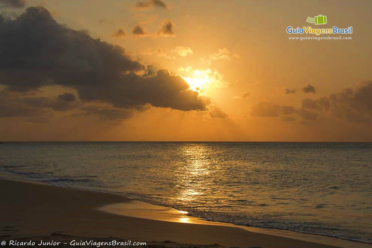 Imagem do Sol alaranjado já se escondendo atrás das nuvens e o mar calmo da Praia da Conceição, em Fernando de Noronha, Pernambuco, Brasil.