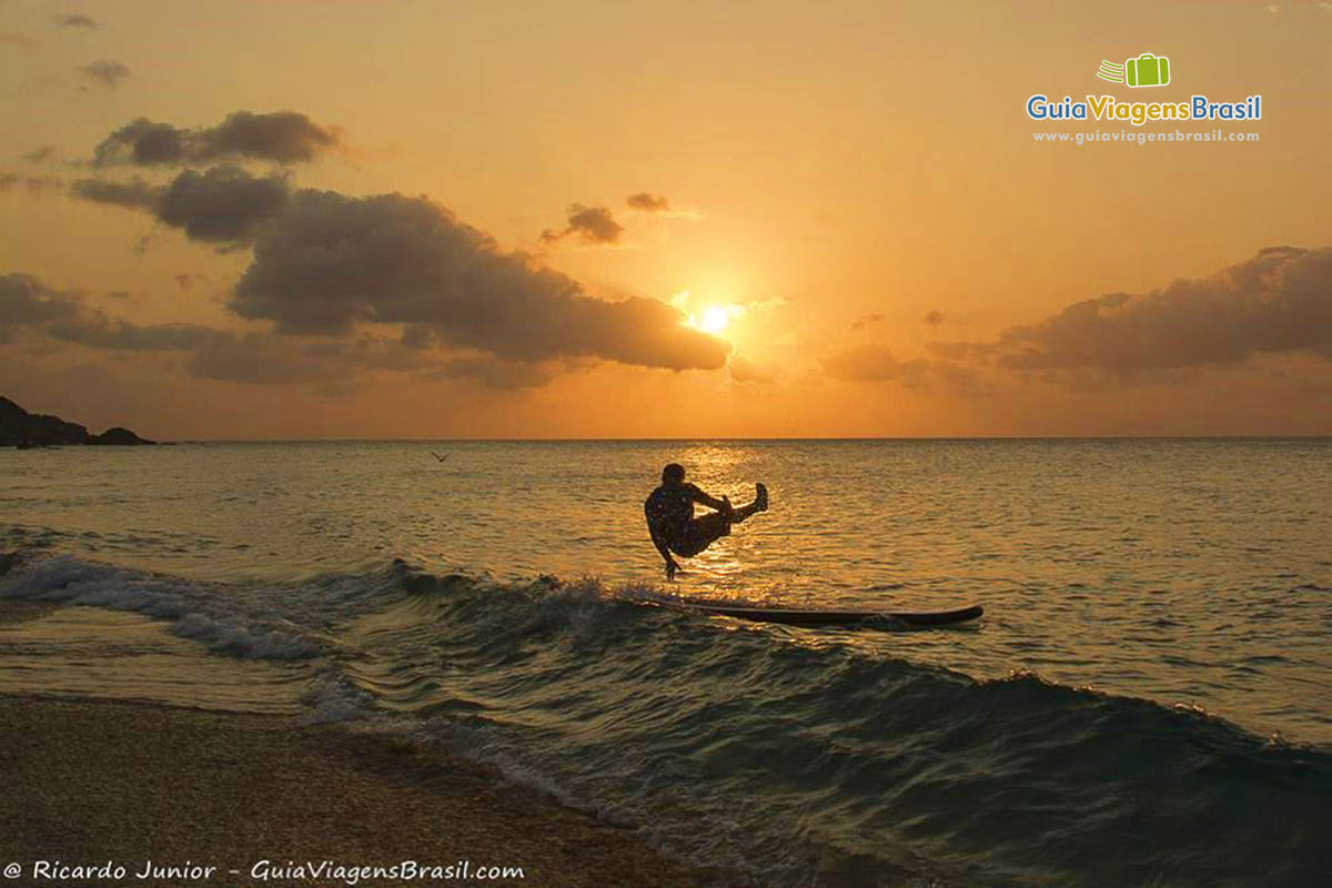 Imagem de turistas nas águas do mar praticando esporte, enquanto o sol já esta se pondo, na Praia da Conceição, em Fernando de Noronha, Pernambuco, Brasil.