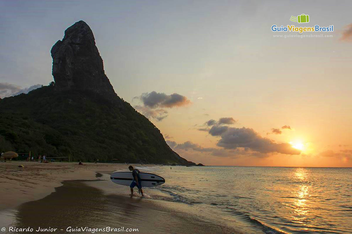 Imagem de turista turista saindo no mar com prancha de stand up e um sol se pondo ao fundo, na Praia da Conceição, em Fernando de Noronha, Pernambuco, Brasil.