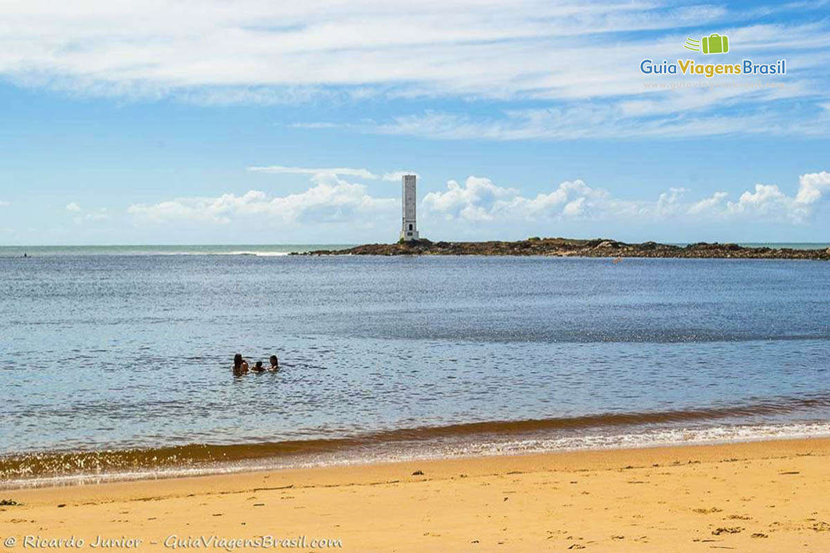 Imagem de crianças brincando na água e ao fundo o farol da bela praia.