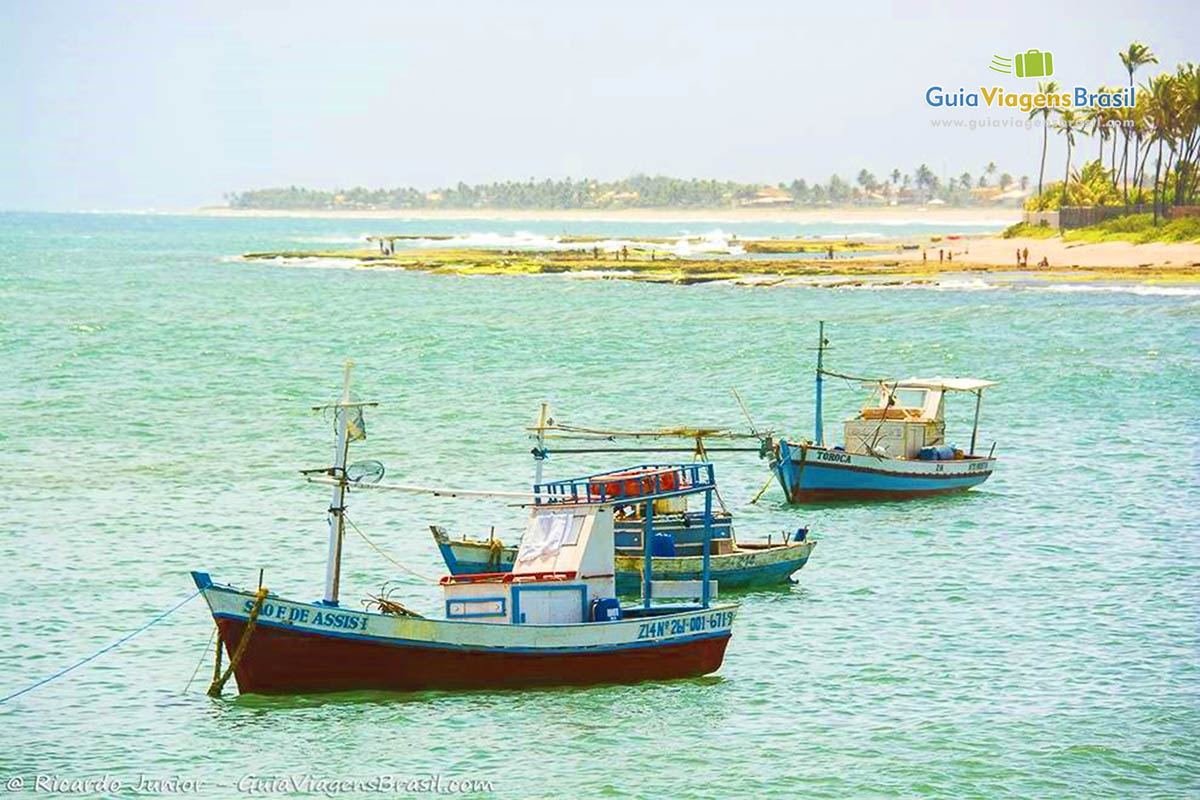 Imagem de três barcos de  pescadores no bar e ao fundo coqueiros e as pedras da Praia de Arembepe.