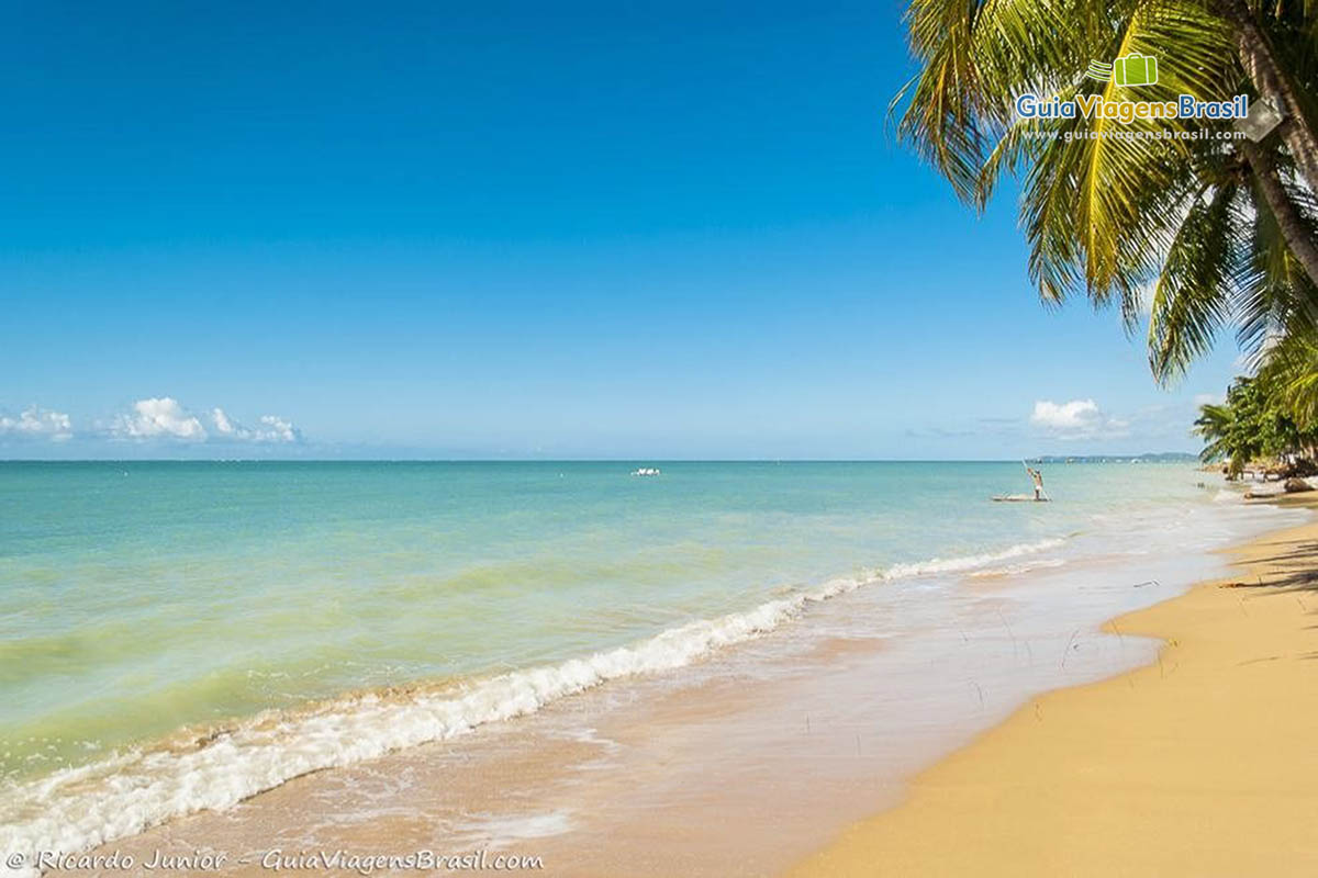 Imagem da praia e ao fundo um pescador em uma jangada em direção ao mar.