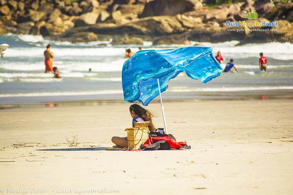 Imagem de uma menina embaixo do guarda-sol se protegendo do sol, na Praia de Fora, na Ilha do Mel, Paraná, Brasil.