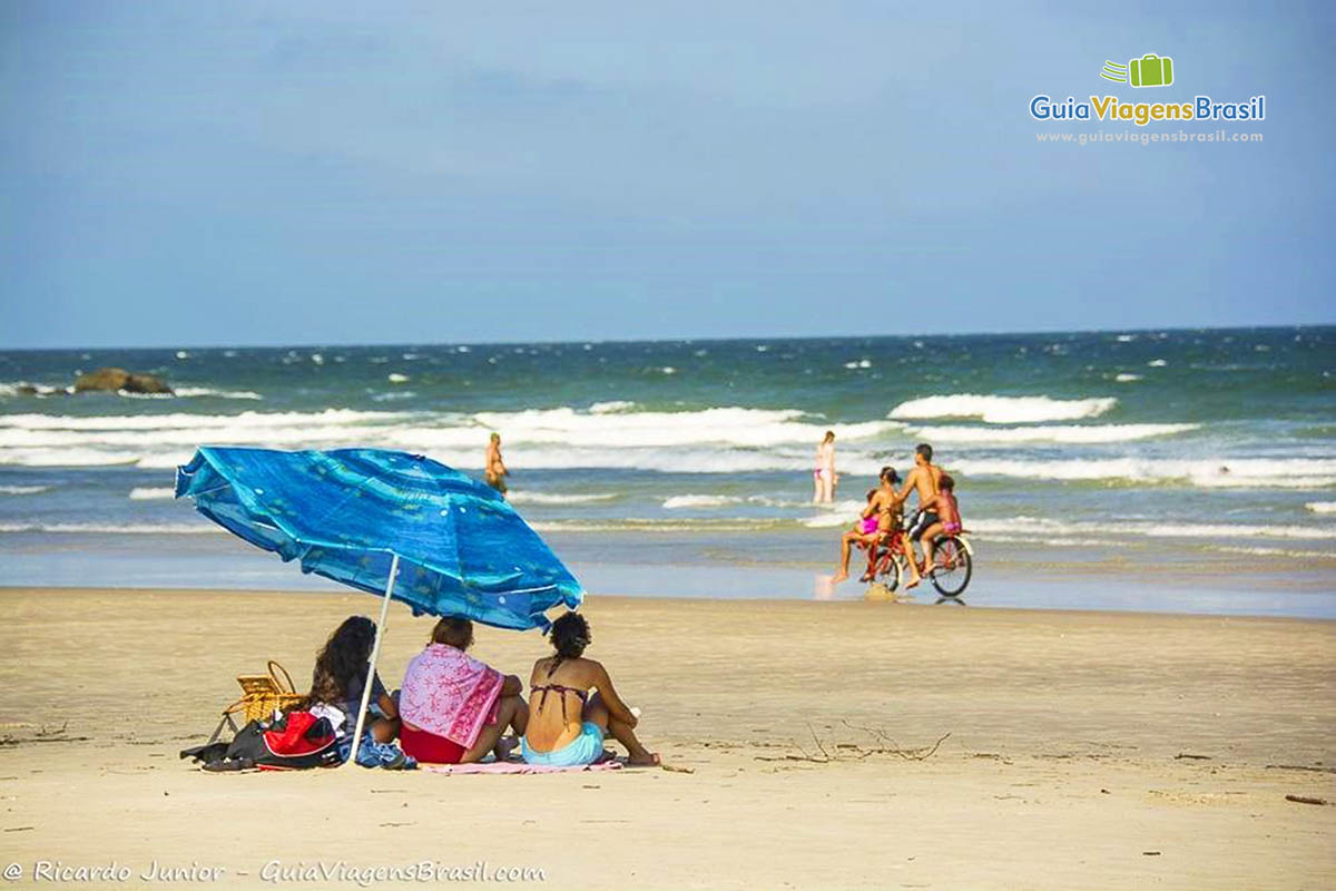 Imagem de três amigas embaixo do guarda sol azul curtindo a paisagem da Praia de Fora, na Ilha do Mel, Paraná, Brasil.