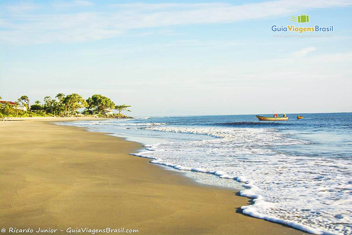 Imagem da Praia de Itapoá, areia dura com espumas brancas na beira do mar e ao fundo um barco, na Praia de Itapoá, em Itapoá, Santa Catarina, Brasil.