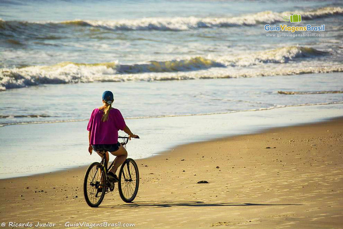 Imagem de uma menina andando de bicicleta na beira do mar, na Praia da Itapoá, em Itapoá, Santa Catarina, Brasil.