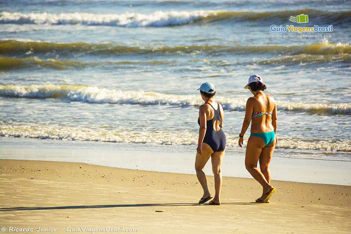 Imagem de duas moças caminhando na beira do mar, na Praia de Itapoá, em Itapoá, Santa Catarina, Brasil.
