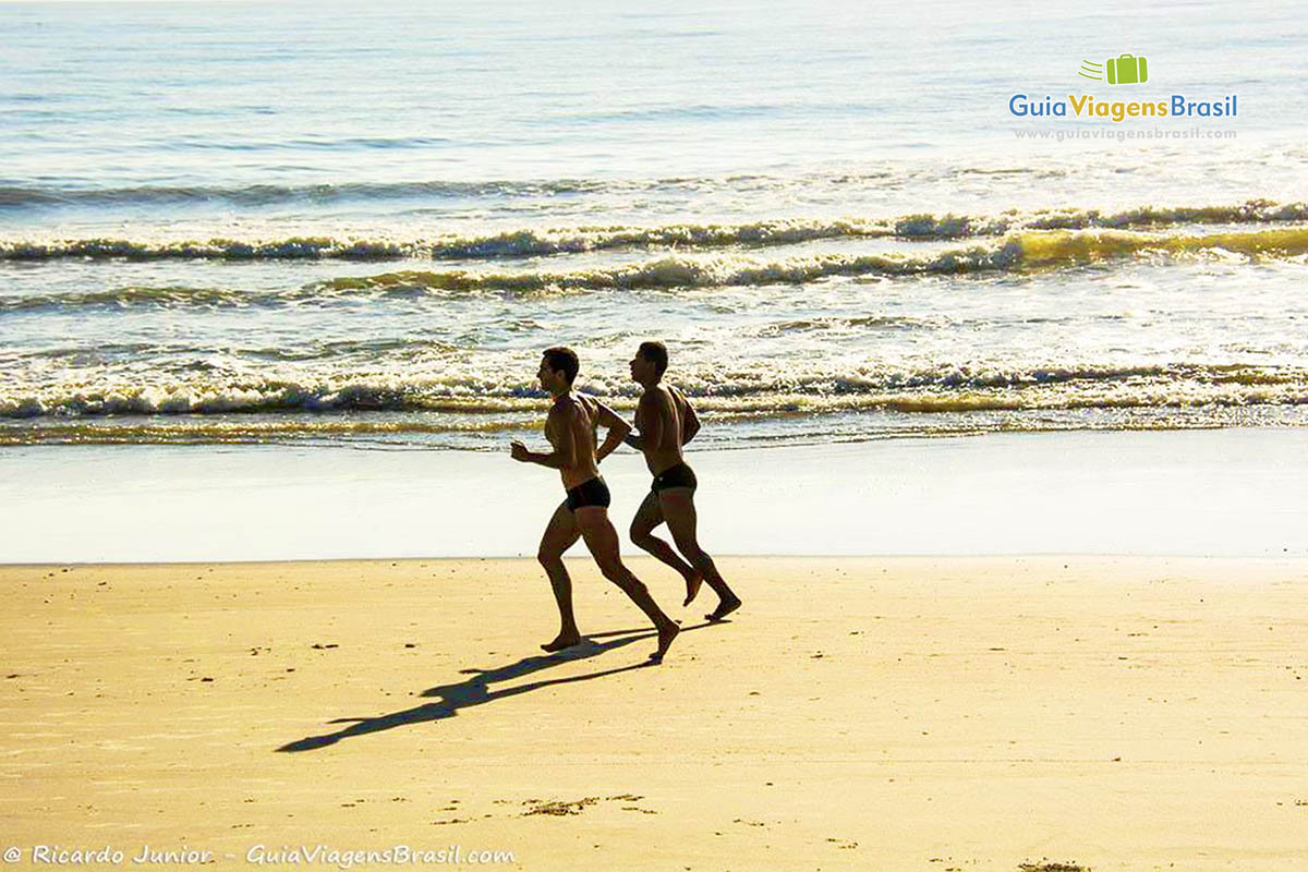 Imagem de dois rapazes correndo na beira do mar, da Praia de Itapoá, em Itapoá, Santa Catarina, Brasil.