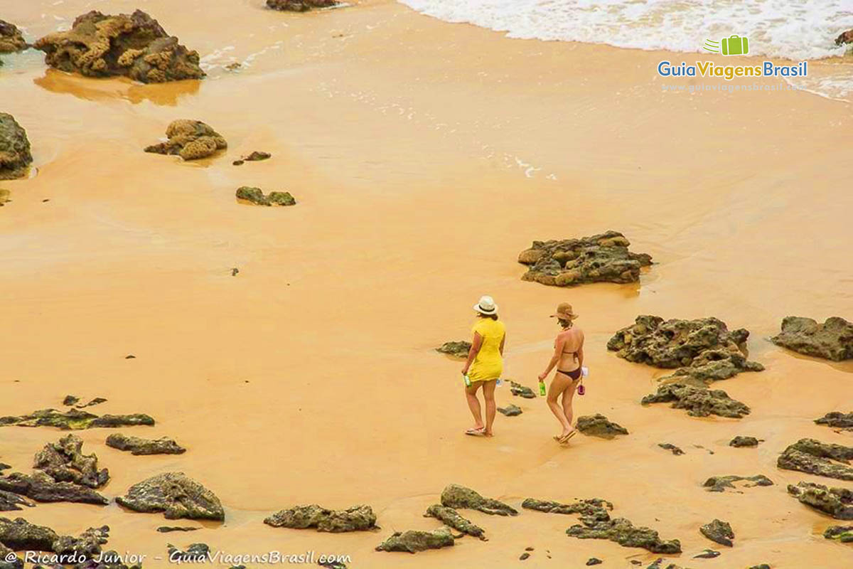 Imagem de duas turistas caminhando na areia entre as pequenas rochas na Praia do Madeiro.