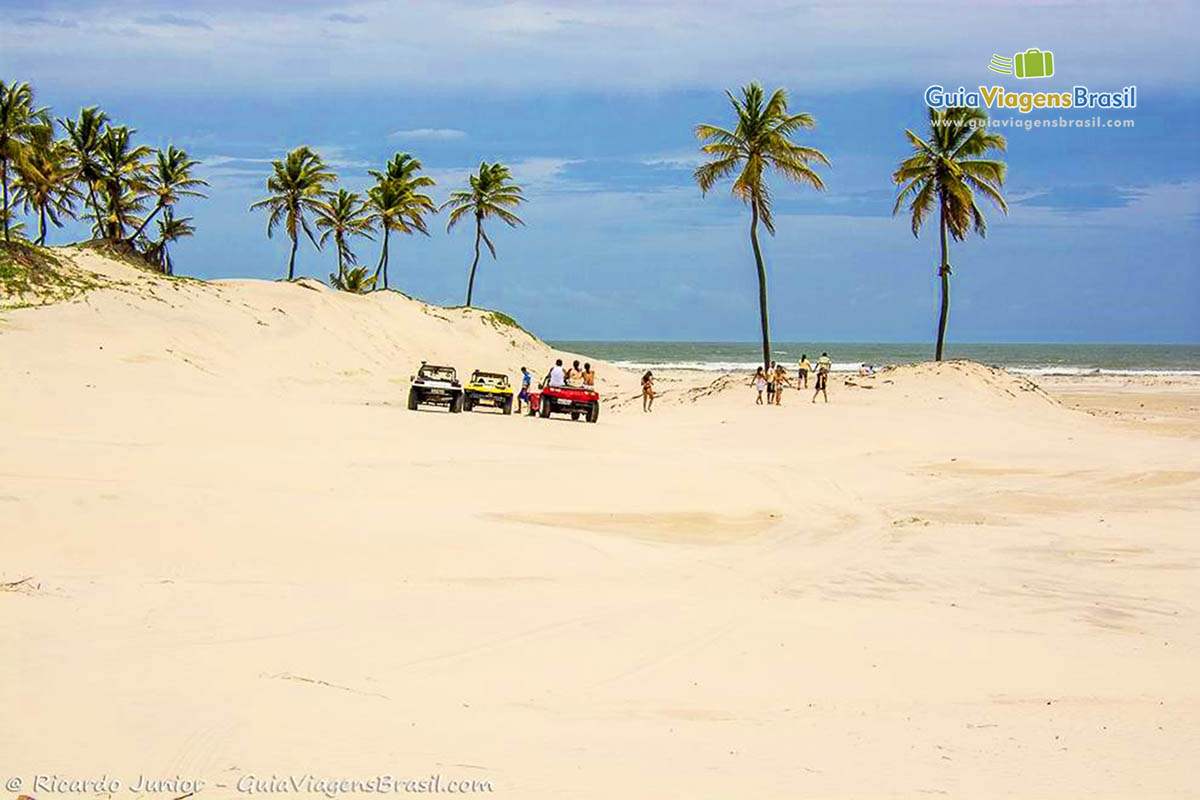 Imagem de turistas caminhando nas dunas e os buggys estacionados.