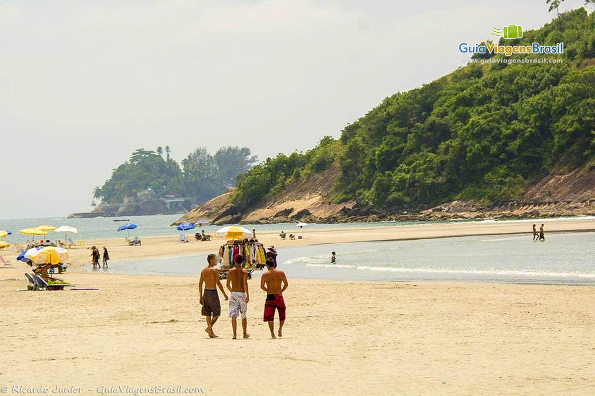 Imagem de três amigos caminhando na areia da praia em direção ao vendedor ambulante.