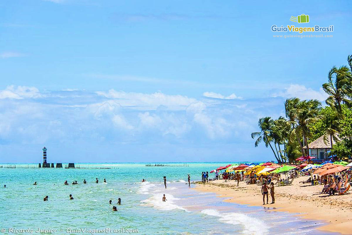 Imagem de turistas nas águas translúcidas e ao fundo o farol da Praia de Ponta Verde em Maceió, Alagoas, Brasil. 