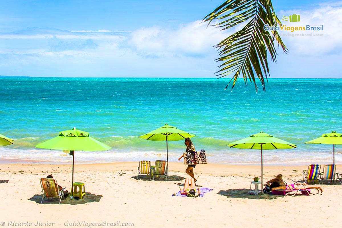 Imagem da Praia de Ponta Verde, com três guarda sol na areia e turistas tomando sol e ao fundo as águas cristalinas do mar, em Maceió, Alagoas, Brasil.