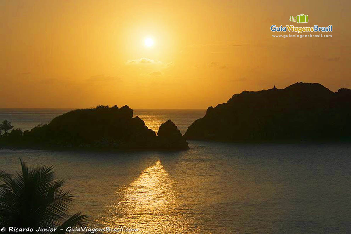 Imagem do alto do pôr do sol, sendo que sol está no meio de duas grandes rochas que estão no mar, em Fernando de Noronha, Pernambuco, Brasil.