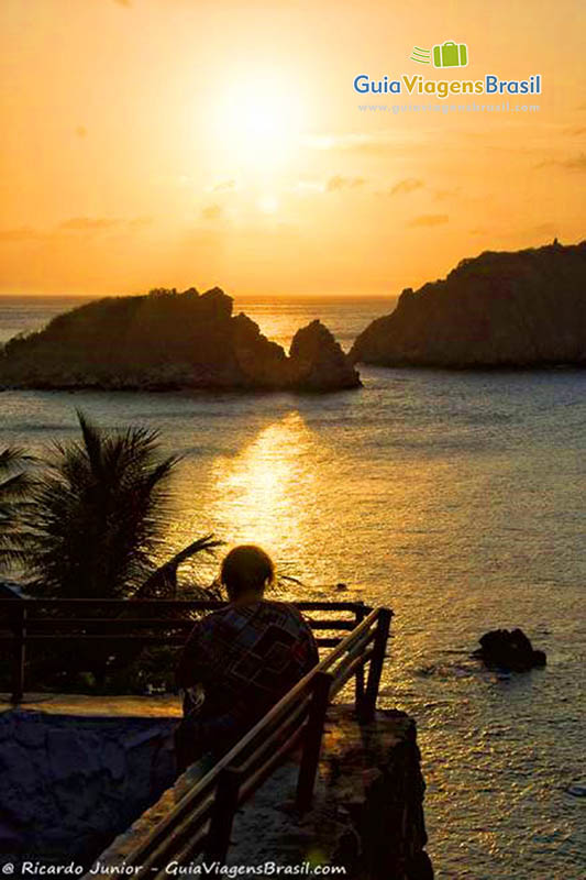 Imagem de uma pessoa sentada no deck, só admirando o pôr do sol, na Praia do Cachorro, em Fernando de Noronha, Pernambuco, Brasil.