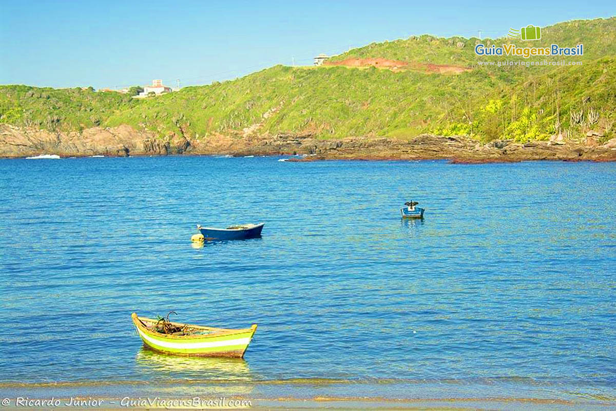 Imagem de barcos na praia e ao fundo morro com duas casas e vegetação rasteira.