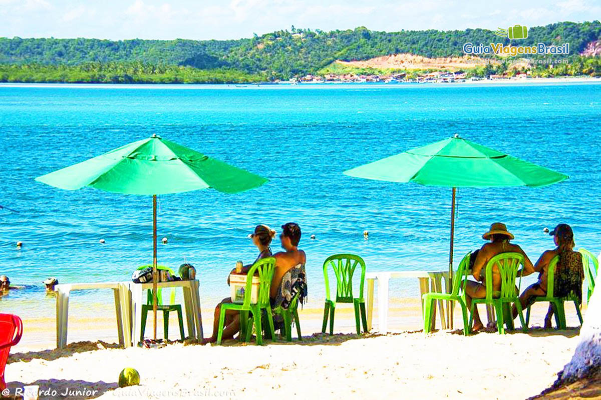 Imagem de turistas sentados com guarda sol na beira da praia admirando a linda Praia do Gunga, em Maceió, Alagoas, Brasil.