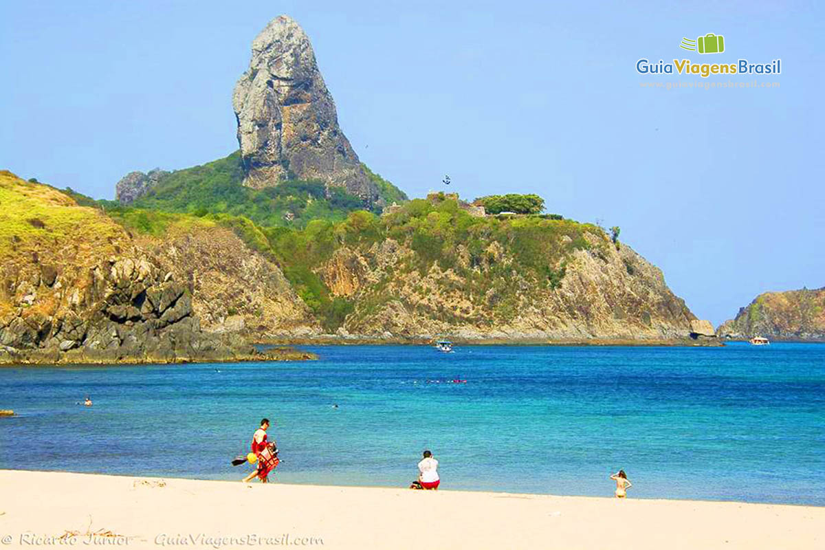 Imagem de turistas nas águas cristalinas e de turista na areia da praia de Porto de Santo Antônio, em Fernando de Noronha, Pernambuco, Brasil.