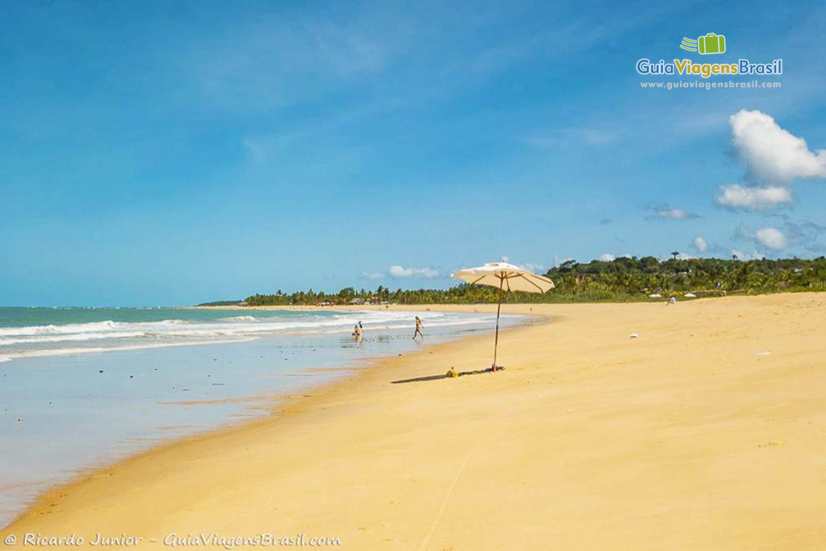 Imagem de um guarda sol branco na areia da praia e alguns turistas no mar, na Praia dos Nativos, em Trancoso.