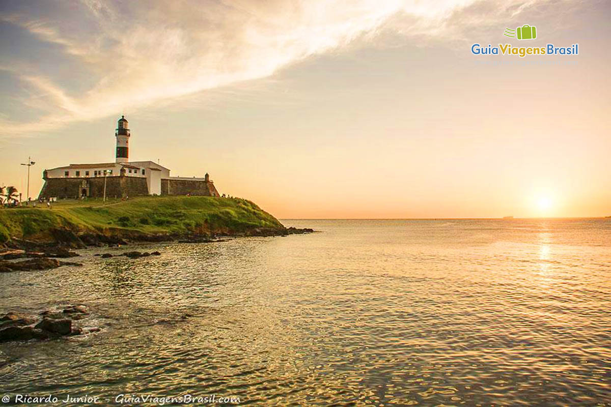 Imagem do lado esquerdo o Farol de lado direito o sol de pondo no horizonte em cima no mar.