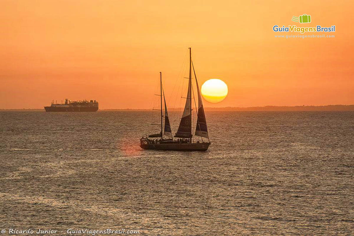 Imagem do por do sol no horizonte, com o céu alaranjado e barco a velas no mar.