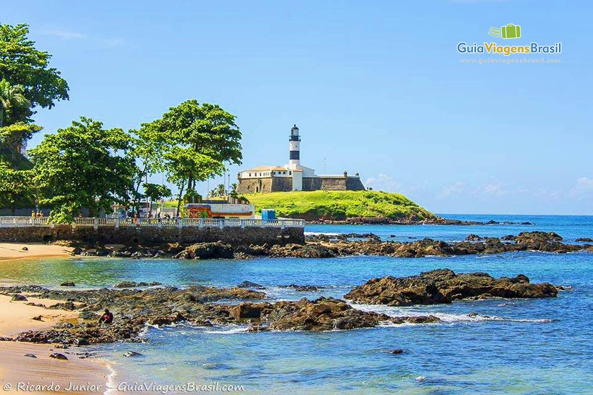 Imagem das águas transparentes e ao fundo árvores e o Forte, na Praia Porto da Barra.