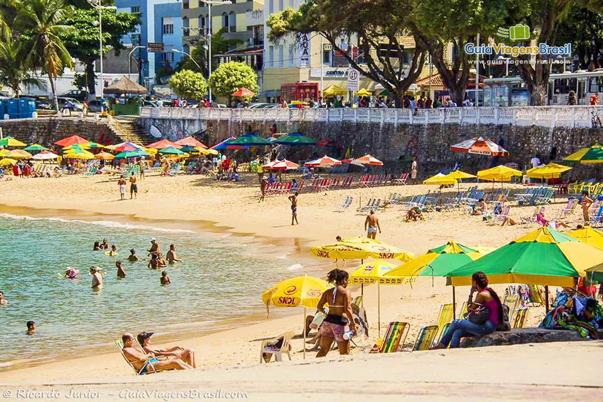 Imagem de turistas na areia com guarda sol, curtindo um dia de praia.