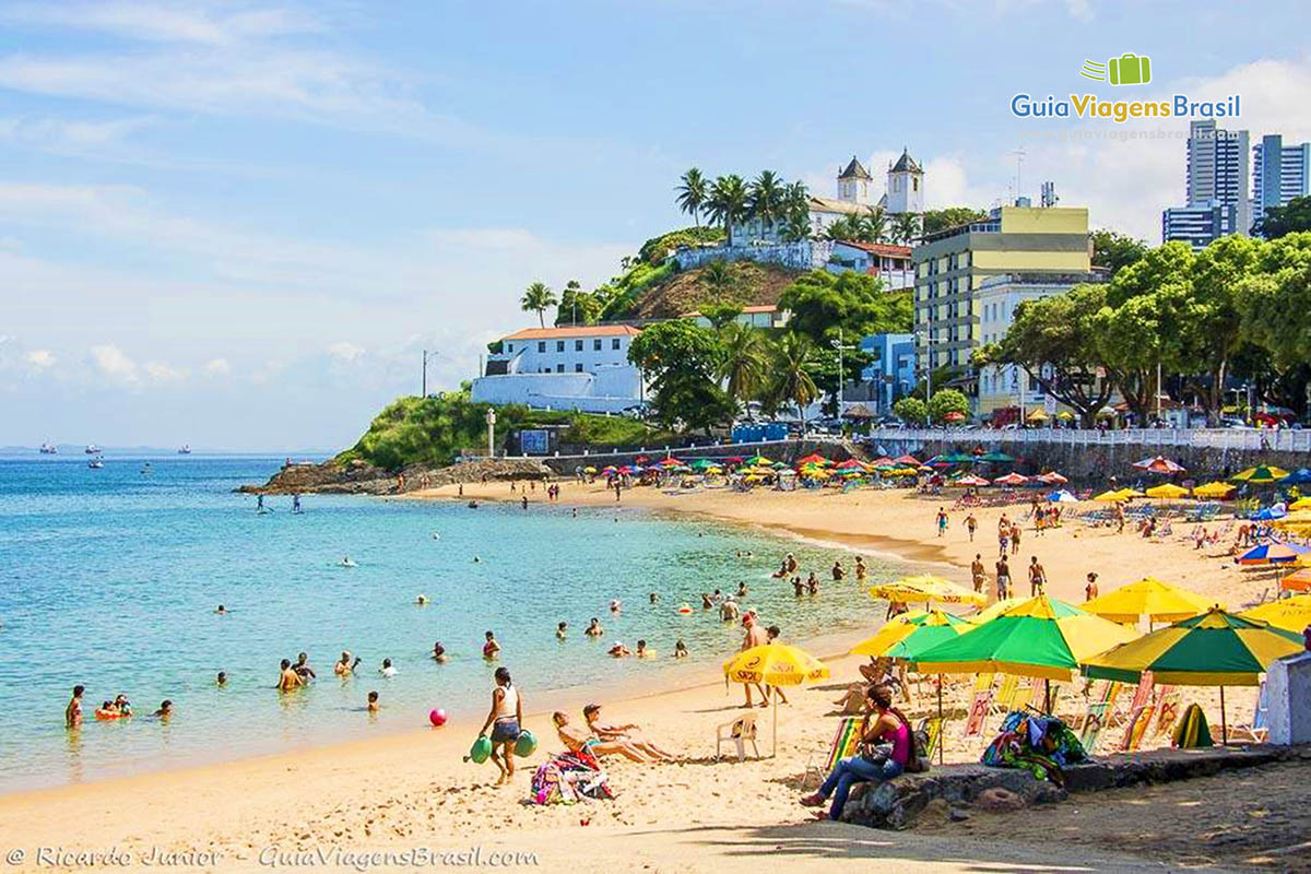 Imagem de toda e extensão da praia, com turistas nas areias e no mar.