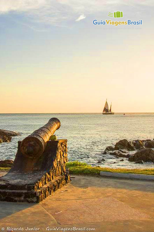 Imagem do canhão e ao fundo o barco a velas no mar, da Praia Porto da Barra.