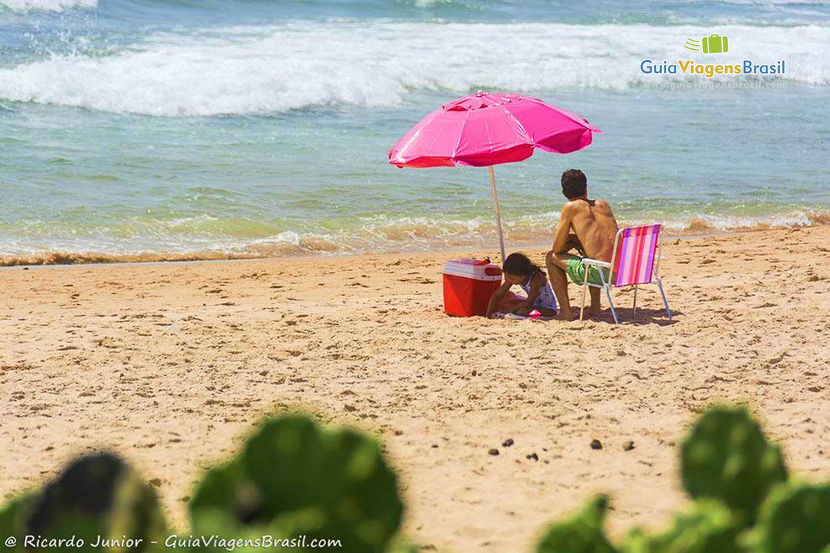 Imagem de turistas sentada em baixo de guarda sol rosa com filha brincando na areia.