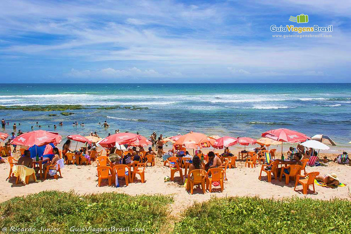 Imagem de vários turistas nas areias e no mar da linda praia.