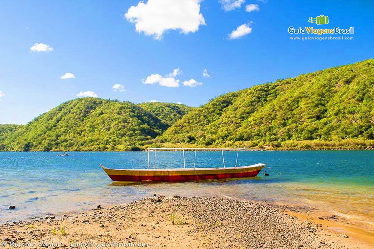 Imagem de lindo barco de pescador parado nas margens do rio, e ao fundo morro com uma vegetação verdinha.