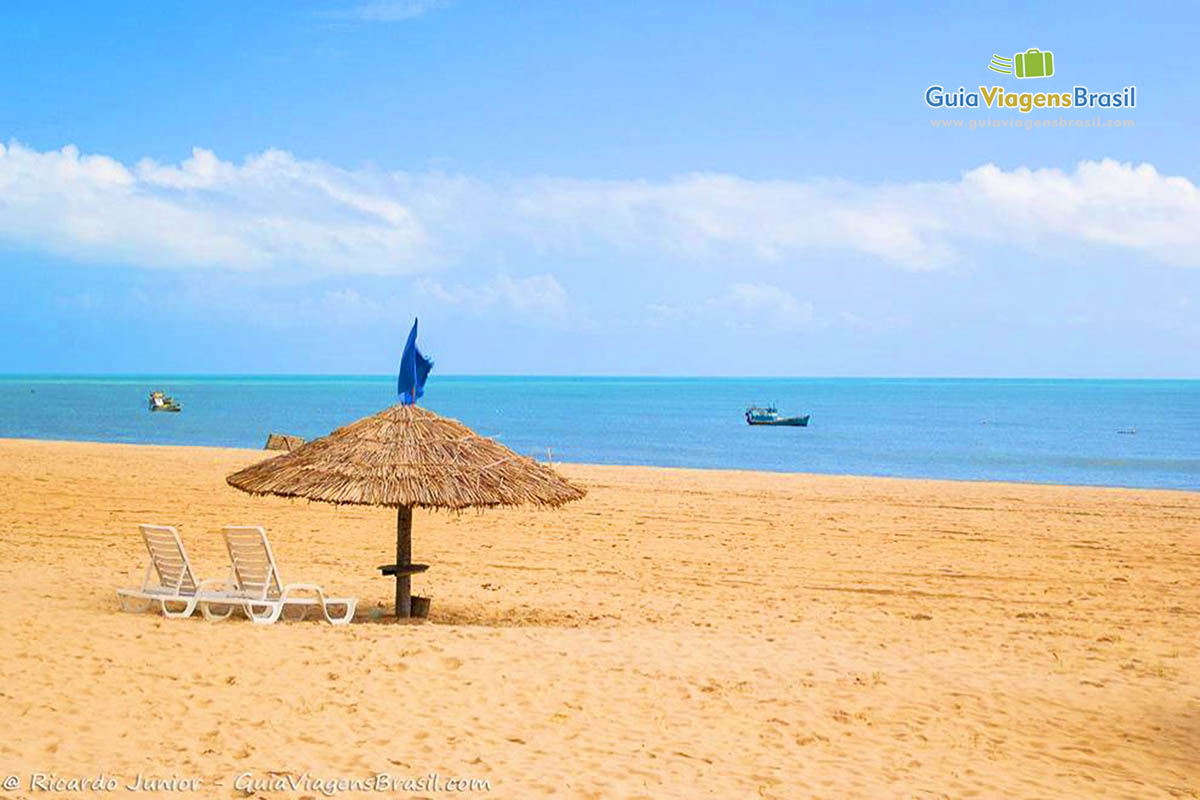 Imagem de um guarda sol de sapê com duas cadeiras e ao fundo mar azul com um barco, na Praia do Maceió.