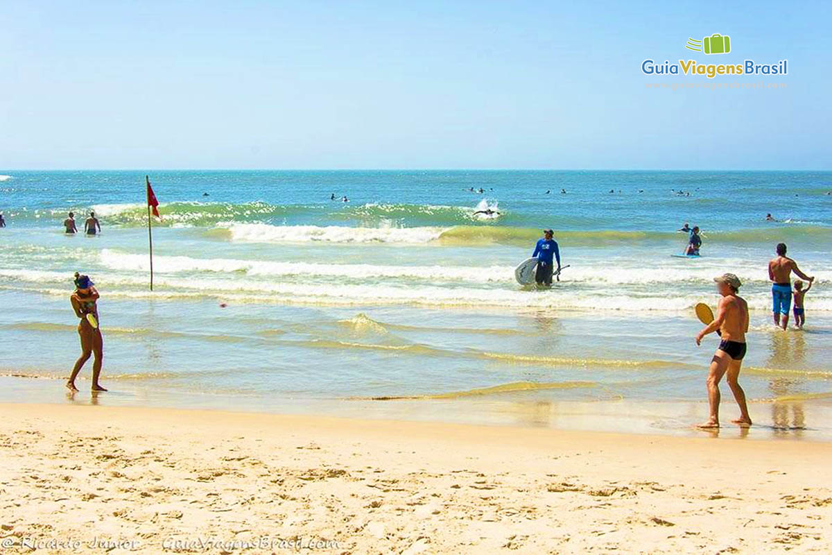 Imagem de duas pessoas jogando frescobol e ao fundo pessoas surfando e praticando stand up paddle.