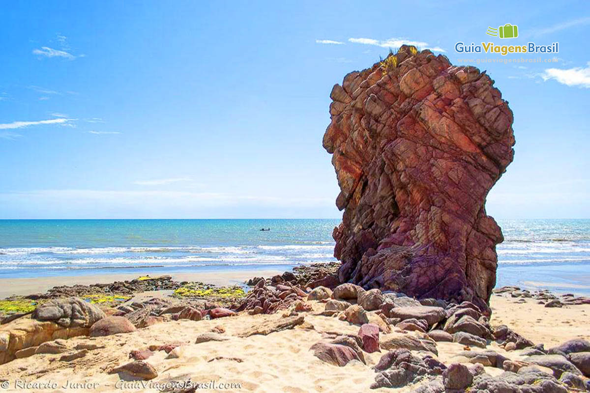 Imagem de grande pedra avermelhada no meio da areia da praia.