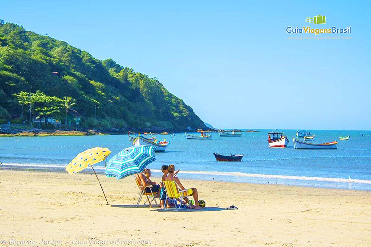 Imagem de crianças e pais sentado na sombra do guarda sol e ao fundo barcos no mar azul da praia.