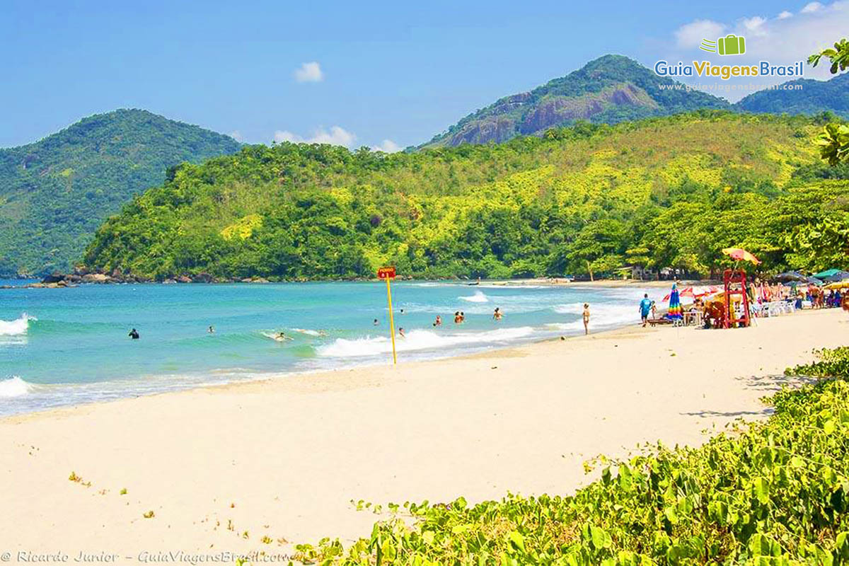 Imagem de turistas em baixo de guarda sol e nas águas da Praia de Castelhanos.