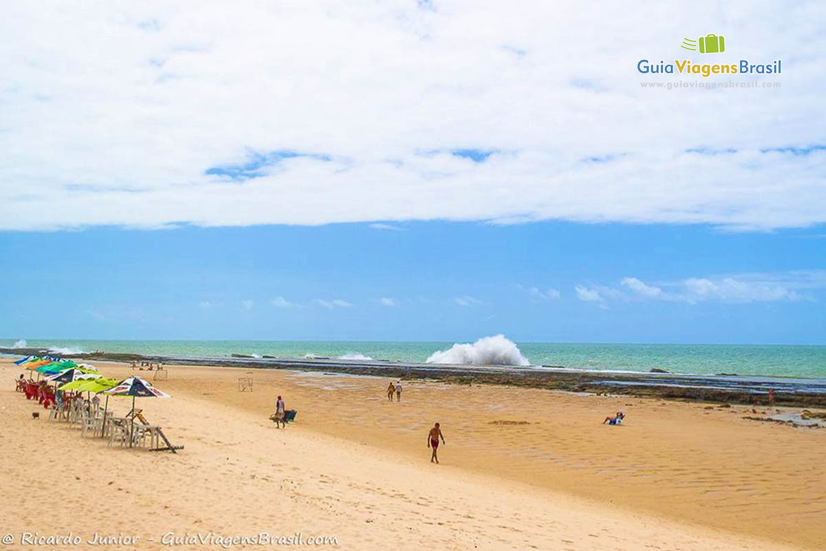 Imagem de guarda sol nas areias e algumas pessoas caminhando na areia da praia.