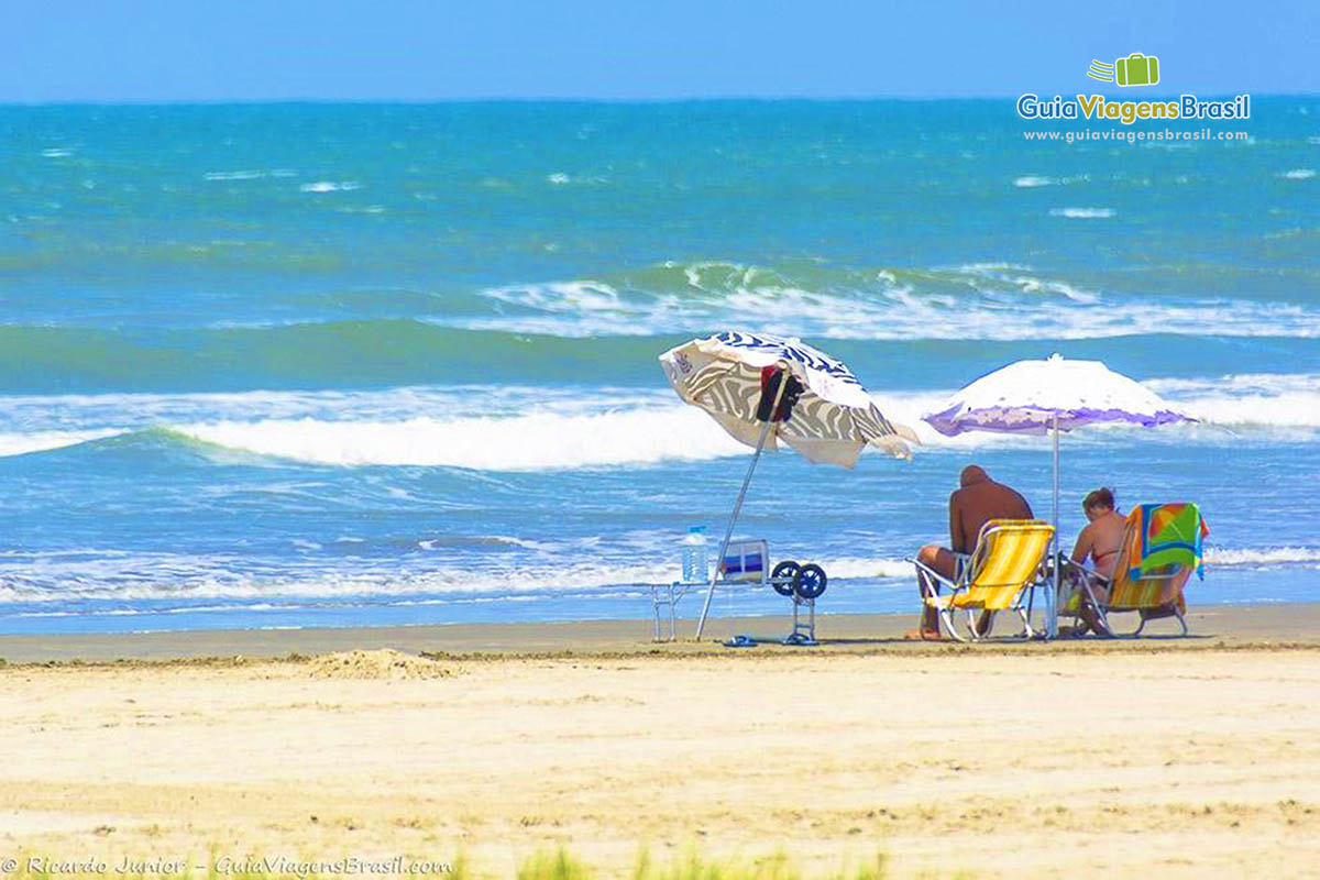 Imagem de casal sentados embaixo de seu guarda sol na praia.