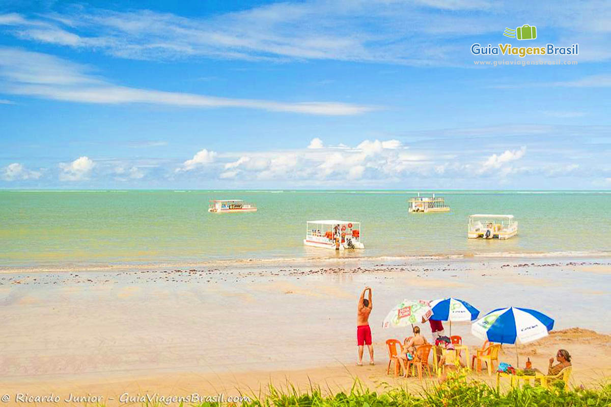 Imagem de turistas nas cadeiras na beira do mar e ao fundo barco de passeio.