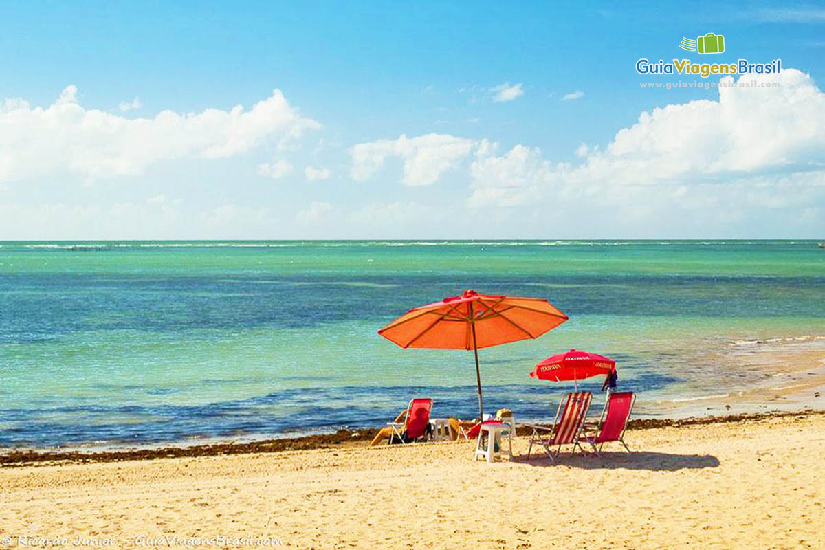 Imagem de casal sentado embaixo de guarda sol vermelho vendo a linda Praia Ponta Verde.