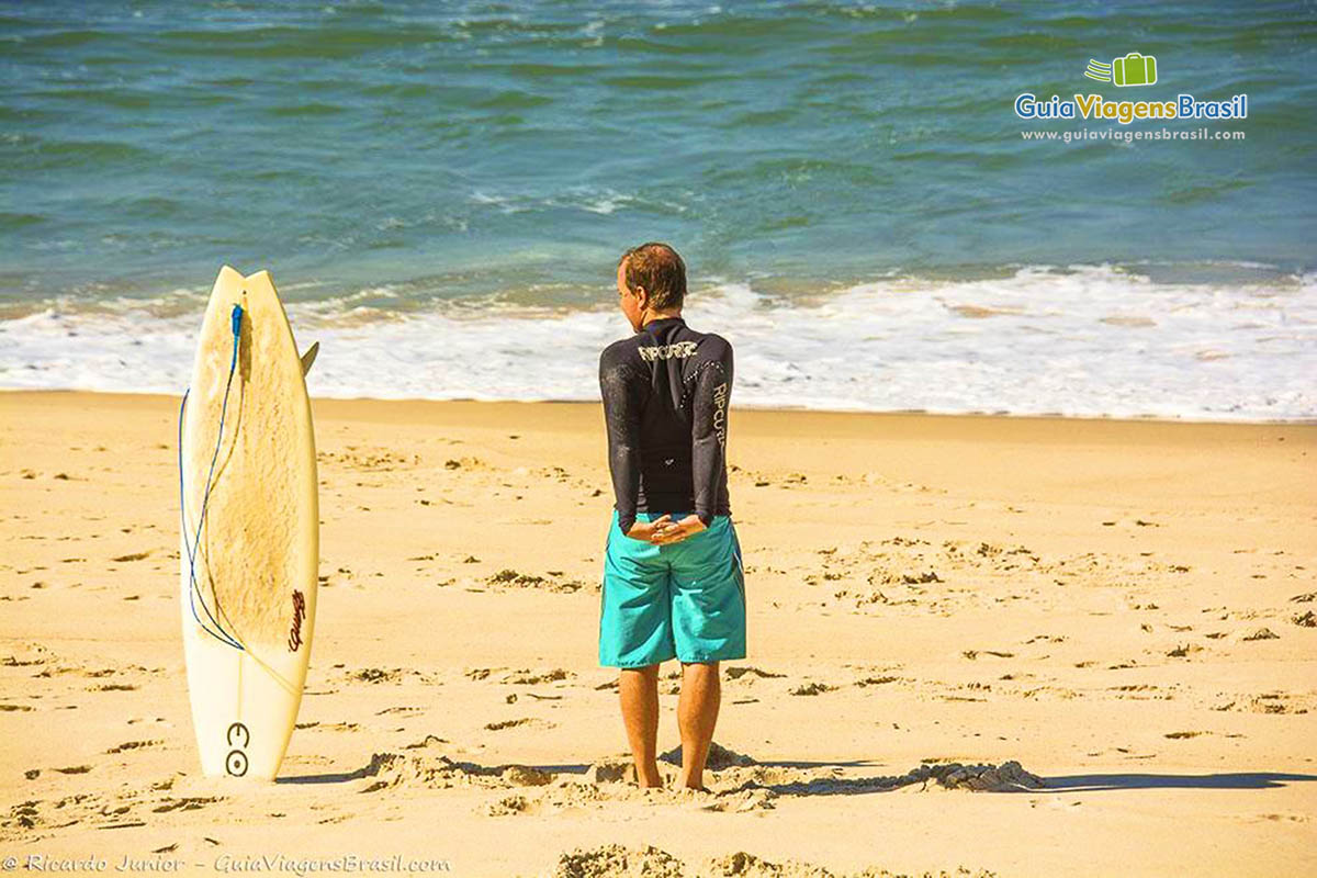 Imagem de uma prancha fincada na areia e surfista ao lado em pé olhando para o mar, dando uma pausa para descanso.