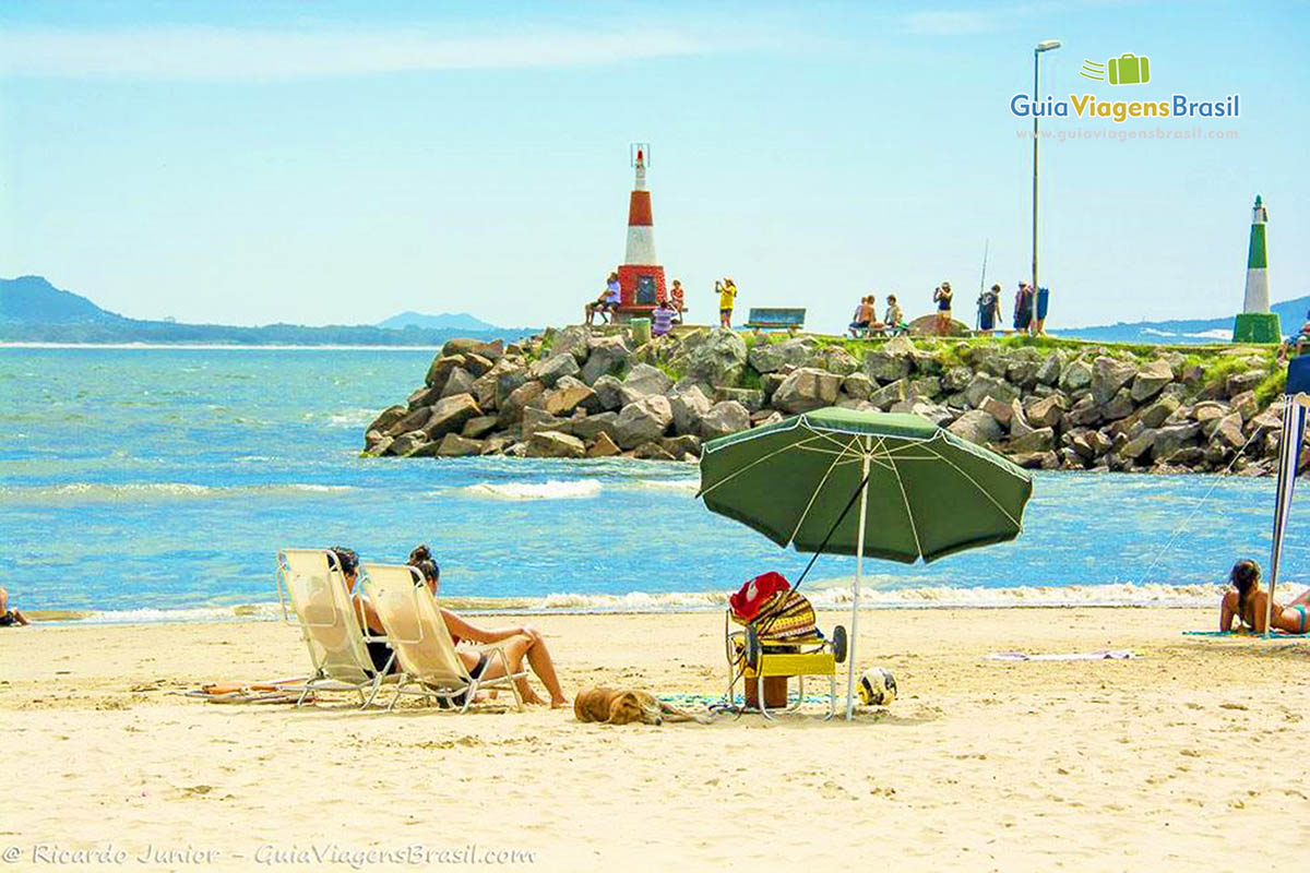 Imagem de casal sentados com cachorro ao lado embaixo do guarda sol e ao fundo turistas nas pedras do farol.